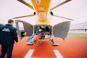 A yellow helicopter is parked with its doors open, revealing an interior filled with medical equipment and a stretcher. A person in a pilot uniform labeled 'PILOTE HELISMUR' is standing nearby on the red landing pad. The surroundings include green grass and a building.
