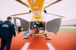 A yellow helicopter is parked with its doors open, revealing an interior filled with medical equipment and a stretcher. A person in a pilot uniform labeled 'PILOTE HELISMUR' is standing nearby on the red landing pad. The surroundings include green grass and a building.