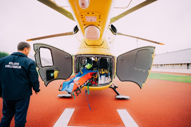 A yellow helicopter is parked with its doors open, revealing an interior filled with medical equipment and a stretcher. A person in a pilot uniform labeled 'PILOTE HELISMUR' is standing nearby on the red landing pad. The surroundings include green grass and a building.