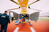 A yellow helicopter is parked with its doors open, revealing an interior filled with medical equipment and a stretcher. A person in a pilot uniform labeled 'PILOTE HELISMUR' is standing nearby on the red landing pad. The surroundings include green grass and a building.