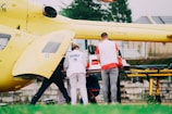 A group of emergency medical personnel are gathered around a yellow helicopter, attending to a patient on a stretcher. The helicopter is equipped for medical emergencies, and the workers are wearing uniforms with 'SAMU' on them. A medical equipment box is placed on the stretcher, indicating a serious or urgent situation.