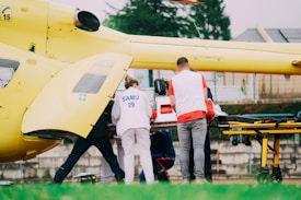 A group of emergency medical personnel are gathered around a yellow helicopter, attending to a patient on a stretcher. The helicopter is equipped for medical emergencies, and the workers are wearing uniforms with 'SAMU' on them. A medical equipment box is placed on the stretcher, indicating a serious or urgent situation.