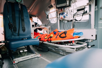 The interior of an emergency medical helicopter is shown with blue seating and a stretcher. Medical equipment and instruments are attached to the walls, alongside an orange survival kit on the stretcher. The atmosphere inside the helicopter is clinical and efficient, reflecting its use for medical emergencies.