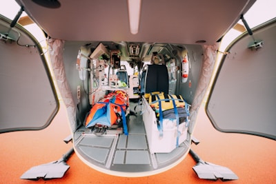 The interior of a helicopter is shown with open doors, revealing a space equipped for medical emergencies. Inside, there is medical equipment, including a stretcher with a colorful red and blue cover and a yellow medical bag. Various control panels and a fire extinguisher are visible, along with seats for staff.