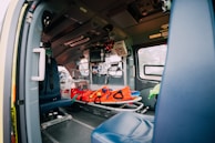 The interior of an emergency vehicle is equipped with medical apparatus, seats, and a stretcher. Various medical equipment and monitors are mounted on the walls, and the space is designed for quick access and functionality.