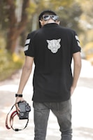 A person wearing sunglasses and a black polo shirt with a geometric animal design on the back is holding a Canon camera by its strap. The scene is set outdoors with blurred greenery in the background, suggesting a casual and relaxed atmosphere.