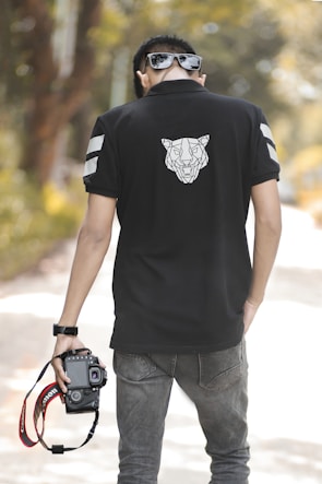 A person wearing sunglasses and a black polo shirt with a geometric animal design on the back is holding a Canon camera by its strap. The scene is set outdoors with blurred greenery in the background, suggesting a casual and relaxed atmosphere.