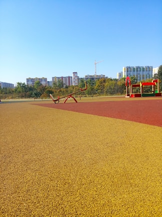 A colorful playground area with red and yellow surfaces, featuring a seesaw and a small play structure. Surrounding the playground are trees, and in the background, a series of modern apartment buildings and a construction crane are visible under a clear blue sky.