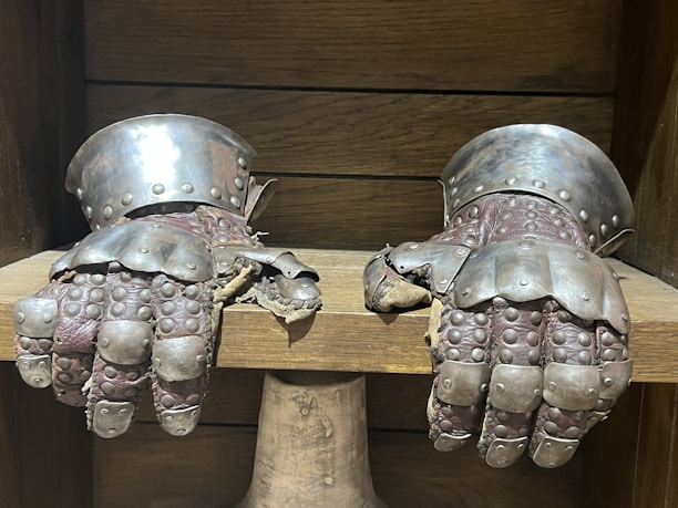A pair of medieval gauntlets made of metal with intricate detailing and studs, resting on a wooden surface. The metal displays a worn texture, and the fingers are articulated with layered plating. The wood behind the gauntlets shows horizontal lines.