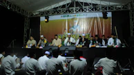 A group of men wearing traditional clothing are seated on a stage under a tent. They appear to be participating in a formal event or ceremony. In front of them, a banner displays a logo and text. The audience, mostly consisting of people sitting on the ground, is visible in the foreground.