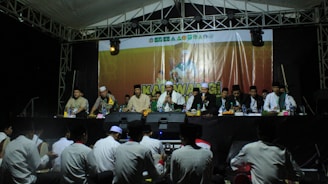 A group of men wearing traditional clothing are seated on a stage under a tent. They appear to be participating in a formal event or ceremony. In front of them, a banner displays a logo and text. The audience, mostly consisting of people sitting on the ground, is visible in the foreground.