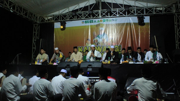 A group of men wearing traditional clothing are seated on a stage under a tent. They appear to be participating in a formal event or ceremony. In front of them, a banner displays a logo and text. The audience, mostly consisting of people sitting on the ground, is visible in the foreground.
