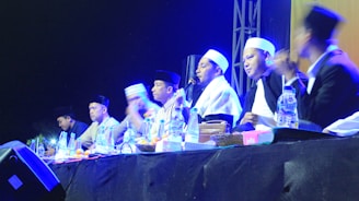 A group of individuals wearing traditional attire and head coverings appear to be engaged in a panel discussion or ceremony. They are seated at a table with various items like bottles of water and fruits in front of them. The setting is dimly lit with blue and yellow stage lights highlighting the participants.