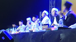 A group of individuals wearing traditional attire and head coverings appear to be engaged in a panel discussion or ceremony. They are seated at a table with various items like bottles of water and fruits in front of them. The setting is dimly lit with blue and yellow stage lights highlighting the participants.