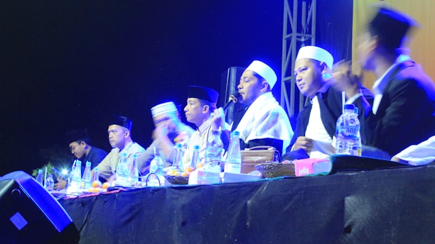 A group of individuals wearing traditional attire and head coverings appear to be engaged in a panel discussion or ceremony. They are seated at a table with various items like bottles of water and fruits in front of them. The setting is dimly lit with blue and yellow stage lights highlighting the participants.