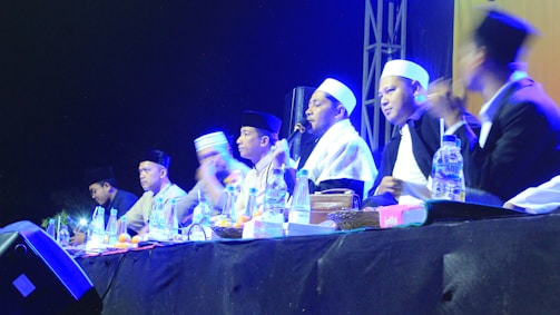 A group of individuals wearing traditional attire and head coverings appear to be engaged in a panel discussion or ceremony. They are seated at a table with various items like bottles of water and fruits in front of them. The setting is dimly lit with blue and yellow stage lights highlighting the participants.