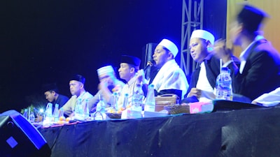 A group of individuals wearing traditional attire and head coverings appear to be engaged in a panel discussion or ceremony. They are seated at a table with various items like bottles of water and fruits in front of them. The setting is dimly lit with blue and yellow stage lights highlighting the participants.