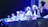 A group of individuals wearing traditional attire and head coverings appear to be engaged in a panel discussion or ceremony. They are seated at a table with various items like bottles of water and fruits in front of them. The setting is dimly lit with blue and yellow stage lights highlighting the participants.