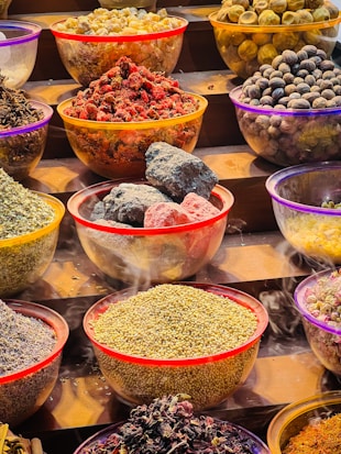Various bowls on a staircase are filled with colorful spices and dried goods. The bowls are transparent with colorful rims, containing items such as dried red fruits, herbs, spices, and stones. Each bowl showcases different vibrant textures and shapes, suggestive of a market or spice display.
