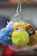 A group of plush keychains featuring small dolls wearing colorful knit hats and fluffy pom-poms. The dolls have round faces with large eyes, and the keychains are held together by metal rings. The background is softly blurred, focusing attention on the vivid, textured dolls.