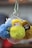 A group of plush keychains featuring small dolls wearing colorful knit hats and fluffy pom-poms. The dolls have round faces with large eyes, and the keychains are held together by metal rings. The background is softly blurred, focusing attention on the vivid, textured dolls.