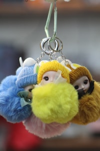 A group of plush keychains featuring small dolls wearing colorful knit hats and fluffy pom-poms. The dolls have round faces with large eyes, and the keychains are held together by metal rings. The background is softly blurred, focusing attention on the vivid, textured dolls.