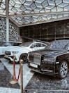 A luxurious indoor setting with a geometric glass ceiling and reflective marble floor. Three high-end luxury cars, including a black and white color scheme, are prominently displayed. A red velvet rope separates the cars from the viewer, suggesting exclusivity.