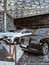 A luxurious indoor setting with a geometric glass ceiling and reflective marble floor. Three high-end luxury cars, including a black and white color scheme, are prominently displayed. A red velvet rope separates the cars from the viewer, suggesting exclusivity.