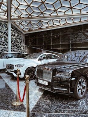 A luxurious indoor setting with a geometric glass ceiling and reflective marble floor. Three high-end luxury cars, including a black and white color scheme, are prominently displayed. A red velvet rope separates the cars from the viewer, suggesting exclusivity.