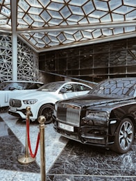 A luxurious indoor setting with a geometric glass ceiling and reflective marble floor. Three high-end luxury cars, including a black and white color scheme, are prominently displayed. A red velvet rope separates the cars from the viewer, suggesting exclusivity.