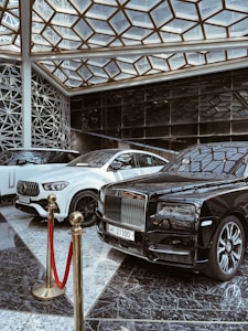 A luxurious indoor setting with a geometric glass ceiling and reflective marble floor. Three high-end luxury cars, including a black and white color scheme, are prominently displayed. A red velvet rope separates the cars from the viewer, suggesting exclusivity.