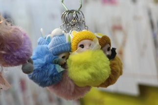 A collection of fluffy keychains with cute doll faces are grouped together. The keychains have knitted caps in various colors like blue, yellow, and brown, which complement the fuzzy textures in matching hues.