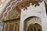 An ornate, decorative interior featuring intricate patterns and luxurious design. The ceiling and walls are adorned with detailed floral and geometric motifs in rich, warm tones. The structure includes lavish gold embellishments and a grand white marble archway with intricate carvings.