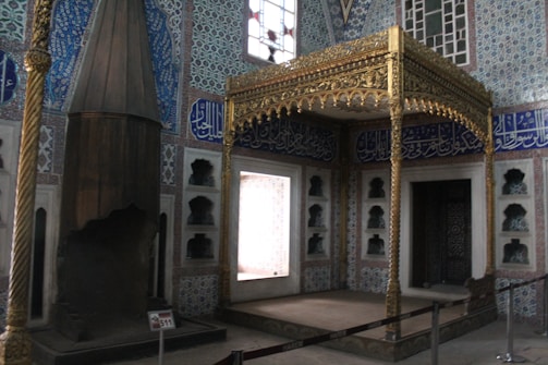 The image depicts an interior with intricate Islamic architectural elements, featuring ornate tile work with geometric and floral patterns. A golden decorative canopy with detailed carvings stands prominently in the scene, and Arabic calligraphy adorns the walls. A tall, dark, conical structure is visible on the left side of the image.