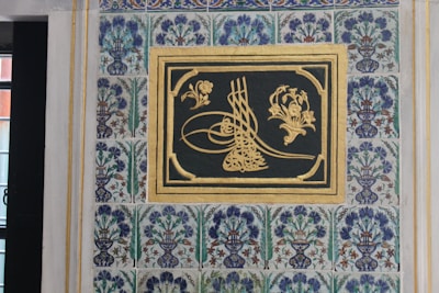 An intricate piece of artwork featuring calligraphy and floral motifs set within a rectangular frame. The background is a tiled wall with repeating patterns of vases holding stylized flowers and leaves in blue, green, and earthy tones. The calligraphy is highlighted with gold, creating an elegant contrast against a dark background.