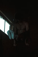 A person is performing gymnastic exercises in a dimly lit room. They are suspended on rings with their upper body and feet visible, wearing white socks. The background has brick walls and a window emitting some light.