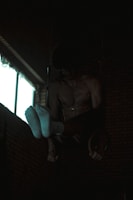 A person is performing gymnastic exercises in a dimly lit room. They are suspended on rings with their upper body and feet visible, wearing white socks. The background has brick walls and a window emitting some light.