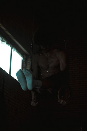 A person is performing gymnastic exercises in a dimly lit room. They are suspended on rings with their upper body and feet visible, wearing white socks. The background has brick walls and a window emitting some light.