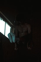 A person is performing gymnastic exercises in a dimly lit room. They are suspended on rings with their upper body and feet visible, wearing white socks. The background has brick walls and a window emitting some light.