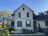 A charming old house with a gabled roof and green shutters, surrounded by a lush garden. The house is painted in light yellow with a dark roof, and there are plants climbing up the walls. The garden features a variety of plants and flowers, with a sunny and bright atmosphere.