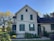A charming old house with a gabled roof and green shutters, surrounded by a lush garden. The house is painted in light yellow with a dark roof, and there are plants climbing up the walls. The garden features a variety of plants and flowers, with a sunny and bright atmosphere.