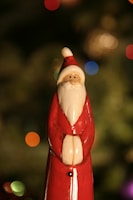 A figurine of Santa Claus with a white beard and wearing a red suit with a pointed hat is placed against a blurred background of multicolored lights and green foliage.