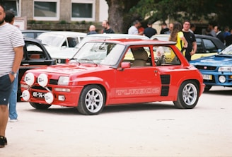 A bright red classic car is parked among several other vehicles at an outdoor car show. Its design is compact with a sporty appearance, featuring decals that read 'TURBO 2' on the side. The car has large rally lights mounted on the front bumper. People are casually standing in the background, some observing the cars.