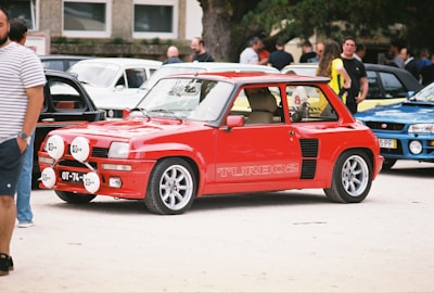 A bright red classic car is parked among several other vehicles at an outdoor car show. Its design is compact with a sporty appearance, featuring decals that read 'TURBO 2' on the side. The car has large rally lights mounted on the front bumper. People are casually standing in the background, some observing the cars.