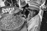 A vendor sits beside a woven basket of beans at an outdoor market, wearing a brimmed hat and a raincoat. Another basket contains wrapped bundles. The scene suggests a small-scale, local marketplace atmosphere.