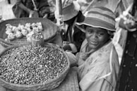 A vendor sits beside a woven basket of beans at an outdoor market, wearing a brimmed hat and a raincoat. Another basket contains wrapped bundles. The scene suggests a small-scale, local marketplace atmosphere.
