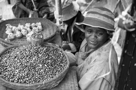 A vendor sits beside a woven basket of beans at an outdoor market, wearing a brimmed hat and a raincoat. Another basket contains wrapped bundles. The scene suggests a small-scale, local marketplace atmosphere.