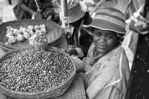 A vendor sits beside a woven basket of beans at an outdoor market, wearing a brimmed hat and a raincoat. Another basket contains wrapped bundles. The scene suggests a small-scale, local marketplace atmosphere.
