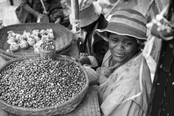 A vendor sits beside a woven basket of beans at an outdoor market, wearing a brimmed hat and a raincoat. Another basket contains wrapped bundles. The scene suggests a small-scale, local marketplace atmosphere.