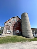 A rustic red wooden barn with a weathered appearance stands next to a tall, cylindrical silo under a clear blue sky. The structure sits on a grassy patch with some small white windows and a gravel path nearby.